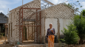 Ibu Fatiha berdiri di depan rumahnya di Desa Ambunten Barat, Sumenep, yang masih dalam proses renovasi. Rumah tersebut sebelumnya berada dalam kondisi memprihatinkan dan nyaris roboh sebelum mendapat bantuan renovasi melalui inisiatif BAZNAS Sumenep. Di atas bangunan itu, bendera Merah Putih tetap berkibar sebagai simbol kecintaan Fatiha terhadap Tanah Air.