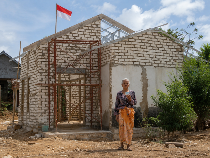 Ibu Fatiha berdiri di depan rumahnya di Desa Ambunten Barat, Sumenep, yang masih dalam proses renovasi. Rumah tersebut sebelumnya berada dalam kondisi memprihatinkan dan nyaris roboh sebelum mendapat bantuan renovasi melalui inisiatif BAZNAS Sumenep. Di atas bangunan itu, bendera Merah Putih tetap berkibar sebagai simbol kecintaan Fatiha terhadap Tanah Air.